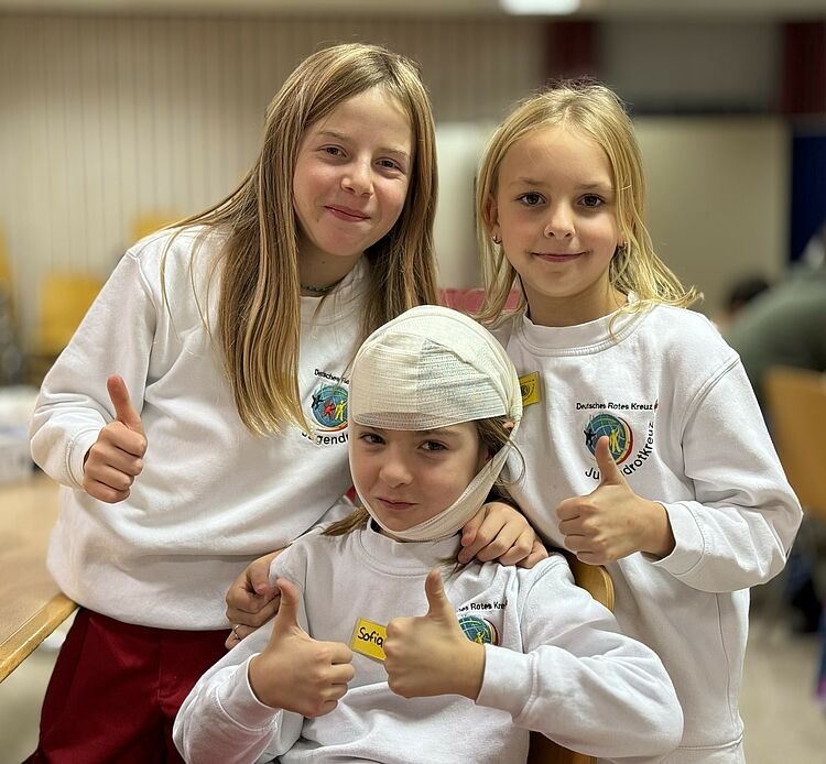 Drei Kinder in JRK-Kleidung lachen in die Kamera. Sie halten den Daumen hoch. Das mittlere Kind trägt einen Kopfverband.