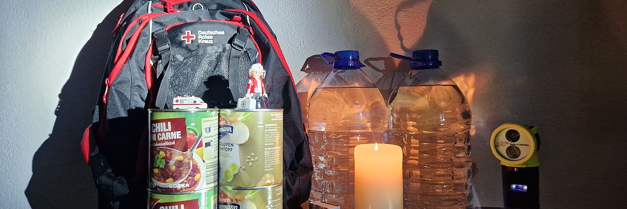 Rucksack, Konserven, Wasserflasche und Taschenlampe stehen im Kerzenschein bereit.