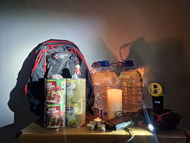 Rucksack, Konserven, Wasserflaschen und Taschenlampe stehen hinter einer brennenden Kerze, bereit für den Gebrauch..