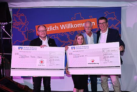 Drei Männer und eine Frau stehen vor einer Fotowand und präsentieren die symbolischen Spendenschecks für die Hauptbegünstigten des Benefiz-GolfCups. Sie lächeln zufrieden. Die beiden Schecks belaufen sich auf jeweils 15.000€.